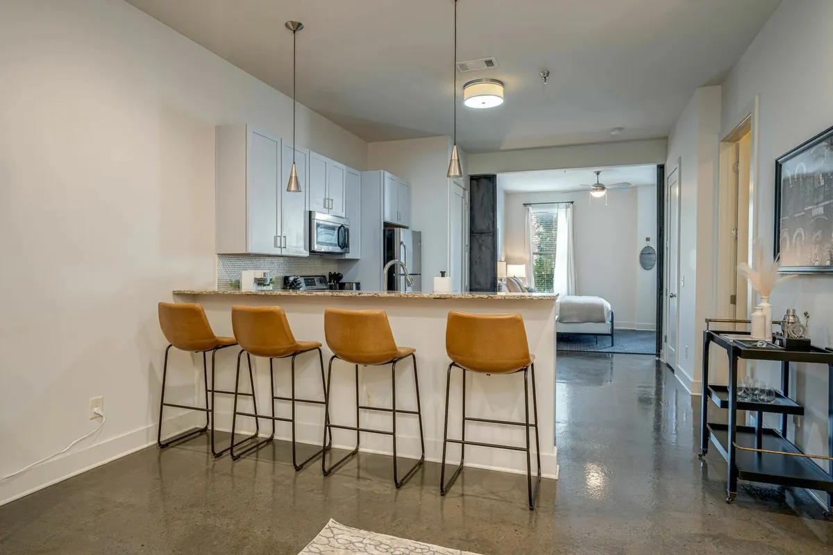 A contemporary kitchen space featuring a central island with bar stools and an open doorway leading into a bedroom. The area is bright, modern, and designed for both cooking and entertaining guests.