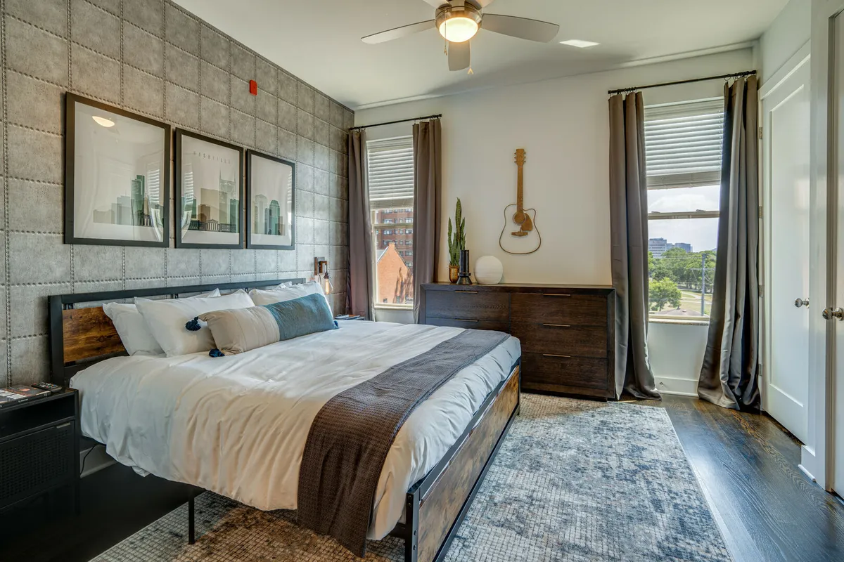 A cozy bedroom with a large bed, bright windows, creative wall art, a guitar resting against the wall, and a dresser. Sunlight floods the space, suggesting relaxation and personal time.