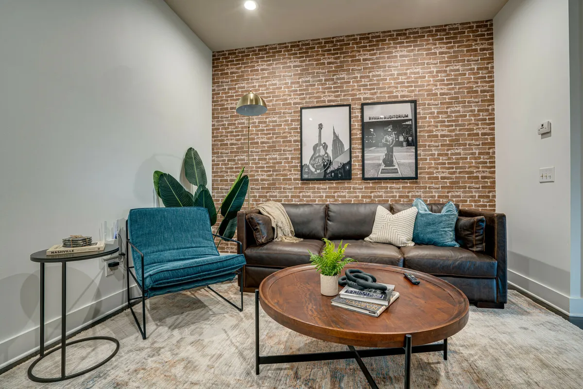 A stylish loft living room with an exposed brick wall, comfortable leather sofa, framed artwork, a green plant, and a wood coffee table. The warm lighting and artful decor create an inviting atmosphere perfect for socializing or quiet relaxation.