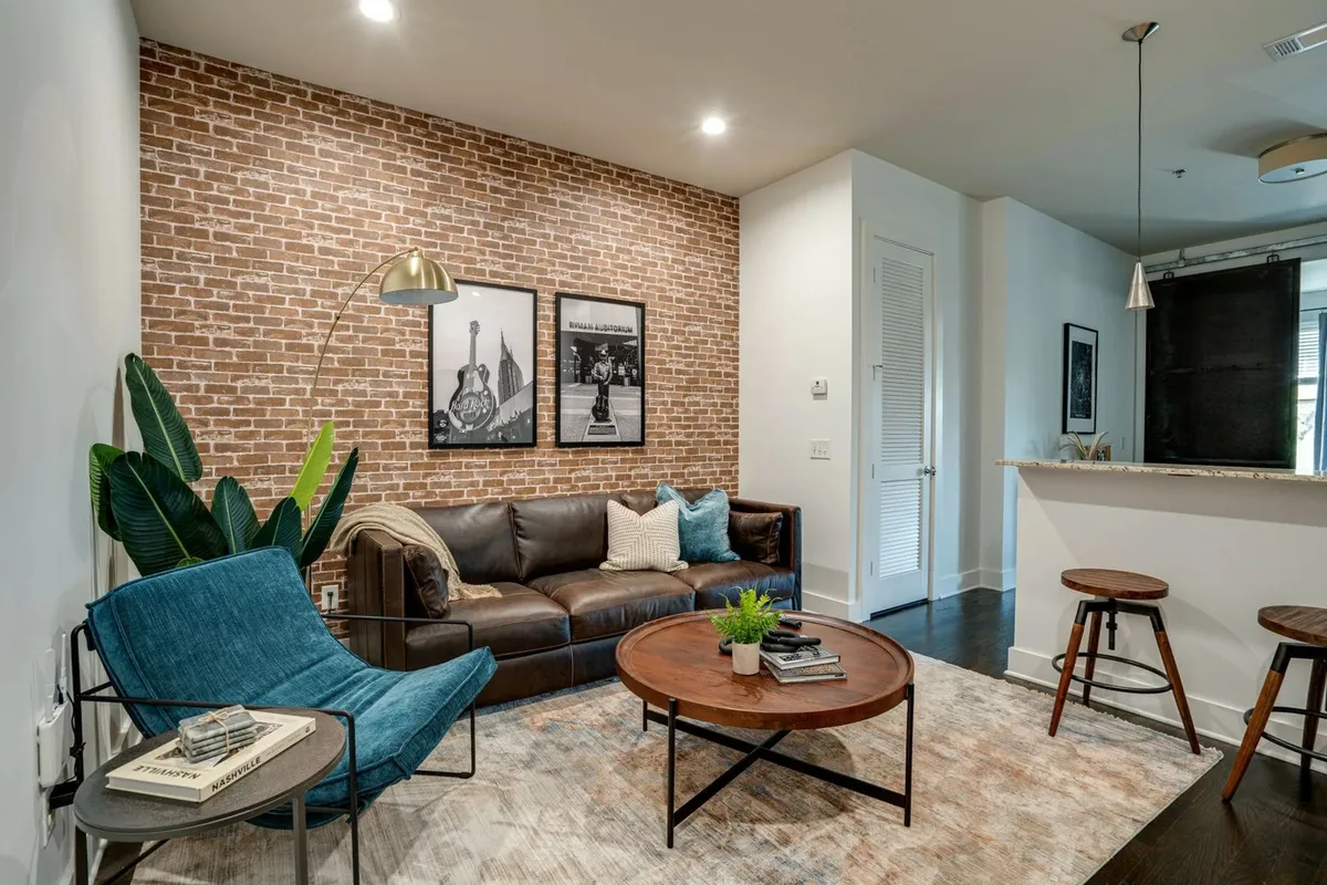 A cozy, contemporary living room with a textured brick accent wall, a chic leather sofa, a coffee table ready for drinks or board games, and a plush blue chair. Wall art adds a touch of personality, creating an inviting space for relaxing or celebrating special moments.