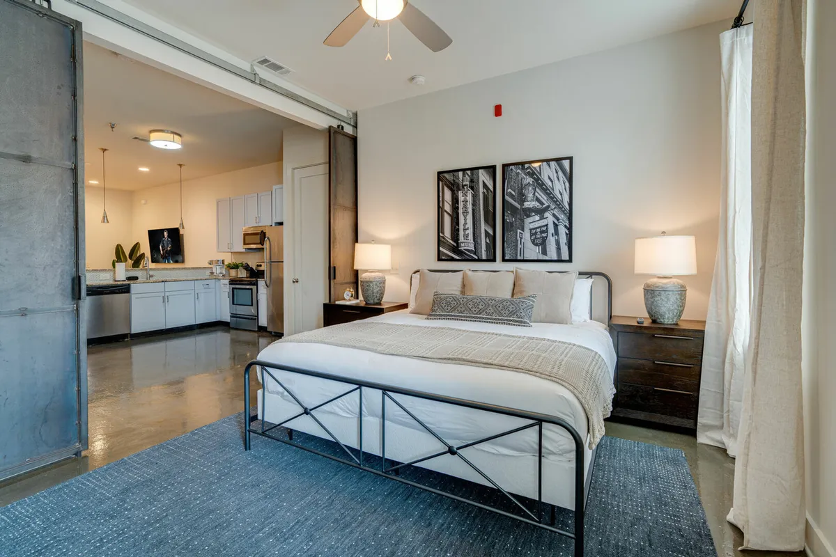 The image showcases a spacious, sunlit bedroom with a stylish bed framed by modern artwork and sleek nightstands, seamlessly opening into a contemporary kitchen area—perfect for both relaxing and enjoying shared meals. The setting is cozy yet sophisticated, embodying the comfort and communal spirit ideal for groups or families visiting Nashville.