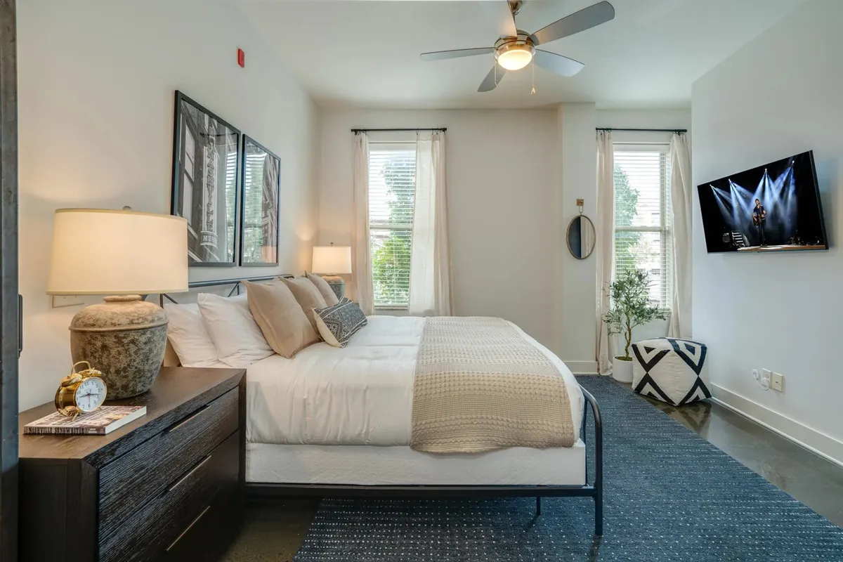 A bedroom featuring a plush king-sized bed with crisp linens, a bedside lamp casting a warm glow, stylish wall art above the headboard, a wall-mounted television for in-room entertainment, and a window that lets in natural daylight.