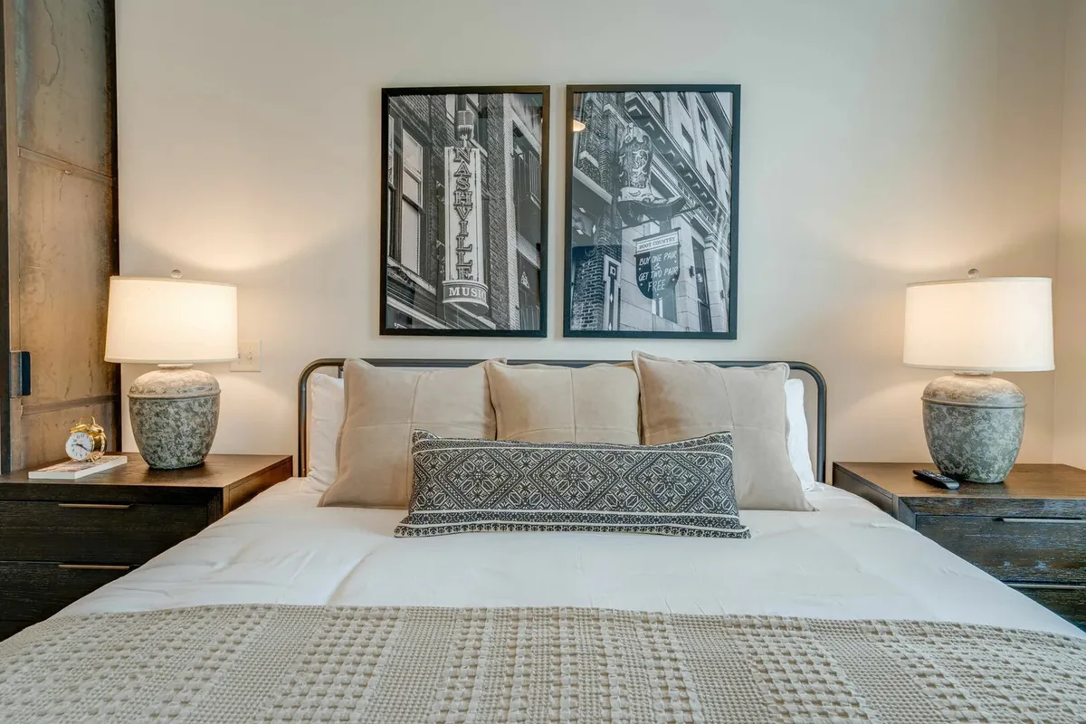 A serene bedroom scene centered on a neatly made bed layered with decorative pillows and soft linens, accompanied by table lamps on either side and elegant black and white photographs framed above the headboard.